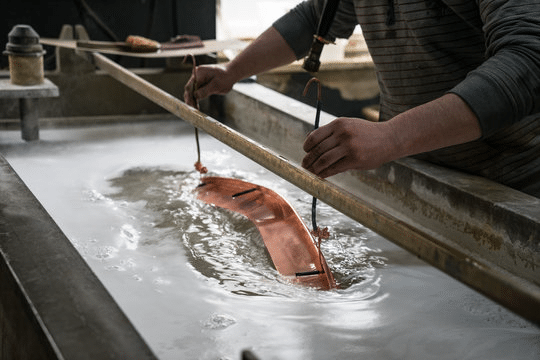 Metalworker uses tongs to lift a curved orange metal casting from a cooling bath in a foundry.